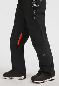 Zwarte sneeuwbroek met een gestructureerde afwerking, voorzien van een oranje binnenvoering, zijzakken met rits en logo-detail. Draagt met zwarte laarzen.