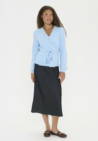 Sieviete ar cirtainiem matiem, tērpta gaišzilā apņemšanas džemperī, melnā midi svārkos un brūnās plakanās kurpēs, stāv pretī vienkāršai baltai fona.