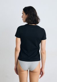 Une femme avec un carré se tient de dos, portant un t-shirt noir et des sous-vêtements gris sur un fond uni.
