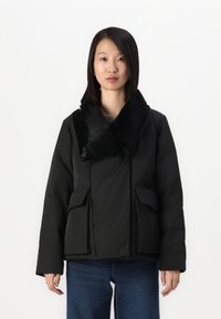 Black puffer jacket with a large, glossy collar, front zip, two side pockets, and a relaxed fit, paired with blue jeans.