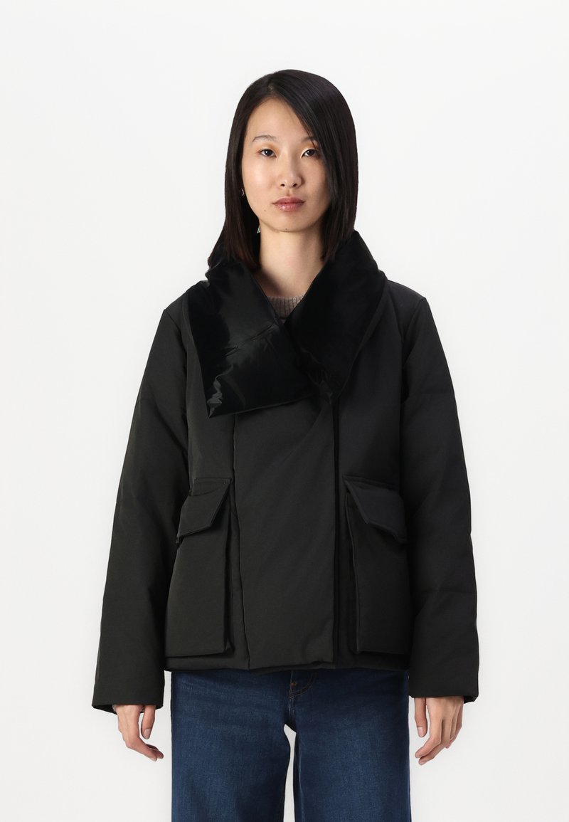 Black puffer jacket with a large, glossy collar, front zip, two side pockets, and a relaxed fit, paired with blue jeans.