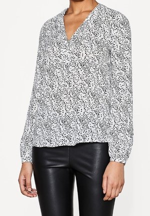 Blouse blanche à manches longues avec motif de points noirs irréguliers, col en V, portée avec un pantalon en cuir noir.
