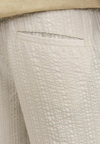 Pantalones de tela texturizada color beige con un patrón sutil en relieve y un bolsillo de ojal. Material suave y ligero con un corte relajado.