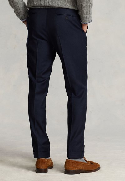 Man wearing dark navy tailored trousers and brown suede loafers, hands in pockets, standing on a light gray floor.