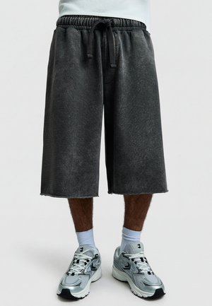 Férfi lábak, fekete, laza, térdig érő rövidnadrágot viselnek, gumis derékpánttal és fekete húzózsinórral, fehér zoknival és ezüst színű sportcipővel.