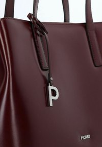 Bordeauxrode leren handtas met een gladde textuur, dubbele handvatten en een zilveren logo-accent. Voorzien van een metalen label met een 'P' bedel.