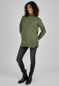 Maglione lavorato a maglia verde oliva con colletto alto, bottoni laterali e orlo a coste, abbinato a jeans neri e stivali neri alla caviglia.