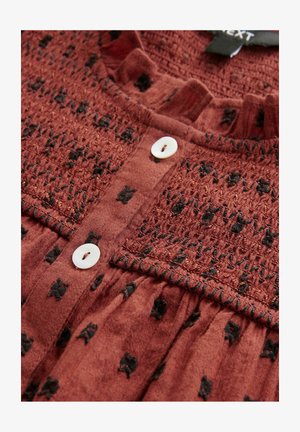 Un capo color ruggine con texture arricciata, caratterizzato da motivi neri. Due bottoni bianchi vicino al collo valorizzano il design.