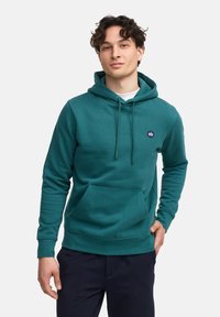 Sudadera verde azulado con un bolsillo tipo canguro, capucha con cordón y un pequeño logo en el pecho. La tela parece suave y texturizada.