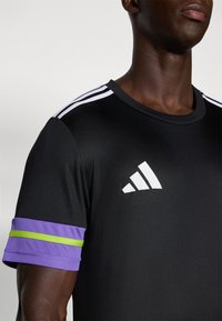 Zwarte sportshirt met drie witte strepen op de schouders, een groot wit logo en paarse mouwen met een groene streepdetails.