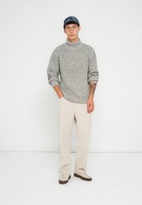 Maglione grigio a collo alto in maglia con maniche arrotolate, abbinato a pantaloni in corduroy beige e sneakers chiare. Il modello indossa un cappellino blu.