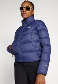 Námornícky modrá puffová bunda s vysokým golierom, prešívaným dizajnom, zipsovým vreckom na hrudi a nastaviteľnými manžetami. Na ľavej hornej strane sa nachádza logo.