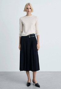 Leichtcremefarbener, gestrickter Pullover mit dezenten Punkten, kombiniert mit einem plissierten, schwarzen Midirock, der mit einem doppelten schwarzen Gürtel und schwarzen Pumps ergänzt wird.