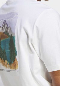 Camiseta blanca de algodón con un gráfico colorido de un paisaje montañoso en la parte posterior, que incluye árboles verdes, un lago azul y foliaje naranja.