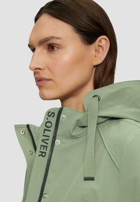 Chaqueta impermeable verde claro con capucha, que presenta un logo negro en la parte delantera, broches plateados y una textura suave. Se aprecia el cierre con cremallera.