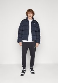 Mornarska pufasta jakna z visokim ovratnikom, zadrgo in logotipom. V kombinaciji s temnimi trenerkami in črno-belimi športnimi copati. Preprost dizajn.