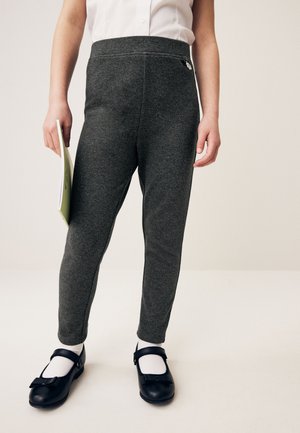 Kind trägt dunkelgraue Hose, weißes Hemd, weiße Socken und schwarze Mary Jane Schuhe, hält in der linken Hand ein hellgrünes Notizbuch.
