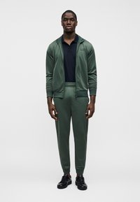 Πράσινη φόρμα με ζακέτα με φερμουάρ και παντελόνι με ελαστική μέση, συνδυασμένη με μαύρο πόλο και μαύρα αθλητικά παπούτσια, με λείο ύφασμα.