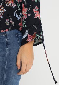 Svart blus med blommönster som har rosa och gröna blommor. Långa ärmar med snörningar vid manschetterna, tillverkad av lätt tyg.