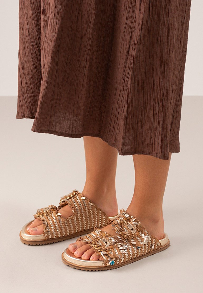 Sandalias trenzadas en tono marrón claro con dobles tiras decorativas que incluyen cuentas y lentejuelas. Suela de goma con detalles texturizados. Combinadas con un vestido arrugado marrón.