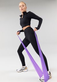 Veste de sport noire zippée et leggings taille haute, tenant une bande de résistance violette, avec des chaussures de course réfléchissantes argentées et dorées.