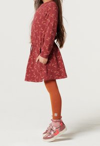 Enfant portant une robe fleurie couleur rouille, des leggings orange et des baskets roses à scratch, debout de profil avec une main dans la poche de la robe.