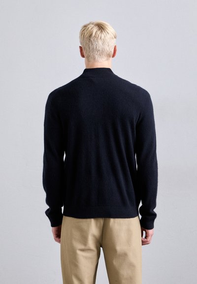 Persoană cu păr blond, îmbrăcată cu un pulover negru cu mânecă lungă și pantaloni bej, întorsă cu spatele împotriva unui fundal gri simplu.