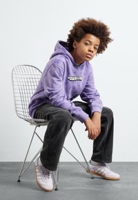 Felpa viola con stampa grafica, pantaloni in corduroy neri e sneakers viola chiaro con strisce bianche, seduto su una sedia di metallo.