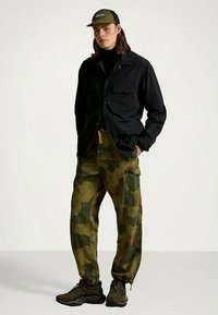 Črna srajca z gumbi, olivno zelene hlače s kamuflažnim vzorcem in temni supergi. Kapa je olivno zelena z logotipom. Priložnostna, funkcionalna obleka.
