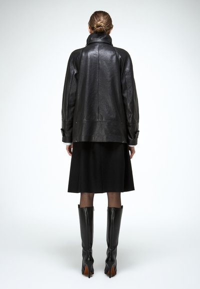 Mujer con el cabello recogido en un moño, vestida con una chaqueta de cuero negra, falda negra, medias transparentes y botas negras de cuero hasta la rodilla, de pie y de espaldas.