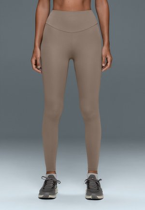 Personne portant des leggings beige taille haute et des chaussures de sport grises, debout sur un fond gris uni.
