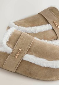 Bruine suède pantoffels met een witte nepbontafwerking, een brede band en metallic "SELF" merknaam aan de zijkant. Zachte textuur en casual ontwerp.