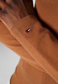 Brązowa koszula z długim rękawem z teksturowanej tkaniny, z małym haftowanym logo w kolorze czerwonym, białym i niebieskim na mankiecie.