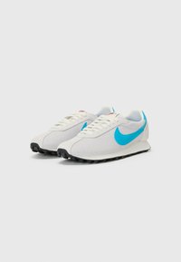 Bela športna obutev s turkiznimi poudarki, mrežasta zgornji del, črn gumijast podplat in bele vezalke. Na strani je logotip Nike.