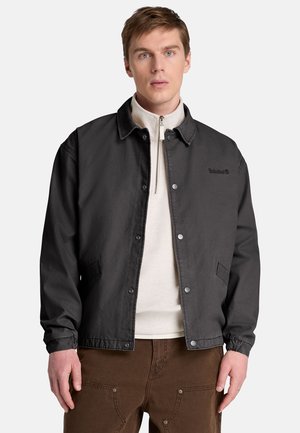 Ung mand iført en sort jakke med knapper over en hvid pullover med lynlås og brune bukser, stående mod en ensartet lys baggrund.
