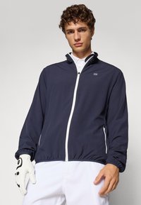 Mornarica jakna na vjetar s bijelim zatvaračem, elastičnim manžetama i ovratnikom. Model nosi golf rukavicu na jednoj ruci i bijelu polo majicu.