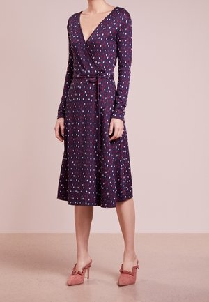 Femme portant une robe portefeuille violette à manches longues avec de petits motifs de cœurs rouges et blancs et des chaussures à talons roses avec des détails à volants.