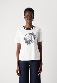 T-shirt bianca in cotone con maniche corte, caratterizzata da un colletto a coste blu navy e una stampa grafica floreale con il nome del brand al centro.