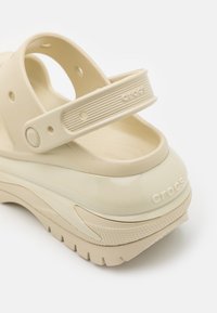 Sandales Crocs beiges avec une semelle épaisse, une bride texturée, une boucle réglable et un logo détaillé sur le talon et la bride. Matériau lisse et durable.