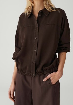 Vrouw draagt een losse, donkerbruine overhemd met knopen en elastische zoom en bijpassende broek, één hand in de zak, neutrale achtergrond.
