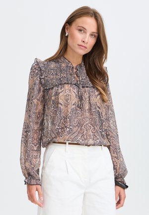 Jeune femme aux longs cheveux porte un chemisier transparent à motif cachemire avec des volants et un pantalon blanc taille haute sur un fond clair uni.
