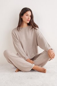 Beige loungewear-set van zachte stof, bestaande uit een loszittend, lang mouwig topje en wijde broekspijpen, met subtiele details aan de zijkant.