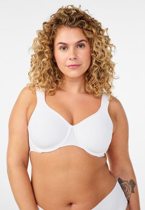 Femme aux cheveux blonds bouclés portant un soutien-gorge et des sous-vêtements blancs, dévoilant un tatouage de rose sur son avant-bras gauche sur un fond blanc uni.