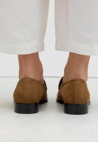 Chaussures en daim marron à talon bas, avec un bout rond et un dos ouvert, posées sur une surface texturée claire, partiellement obscurcies par un pantalon blanc.