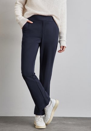 Osoba nosząca granatowe spodnie o prostych nogawkach, białe trampki i kremowy sweter z fakturą, stojąca z jedną ręką w kieszeni na szarym tle.