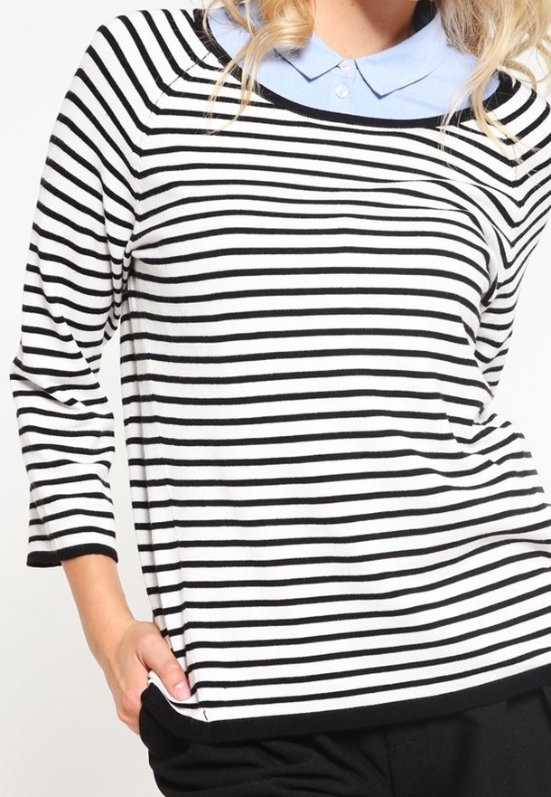Femme portant un chemisier à manches longues à rayures horizontales noires et blanches par-dessus une chemise à col bleu clair, avec une main dans la poche d'un pantalon noir.