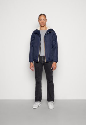 Giacca puffer blu navy con cappuccio foderato in crema testurizzato, chiusura con zip e dettagli in oro, abbinata a un maglione grigio e jeans scuri.