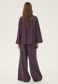 Abbigliamento composto da due pezzi in morbido tessuto di colore viola scuro. La maglia a maniche lunghe ha un orlo arrotondato e i pantaloni a gamba larga scendono fino a terra. Texture liscia.