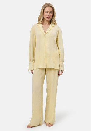 Mujer de pie descalza con una camisa holgada de manga larga color amarillo pálido abotonada y pantalones anchos a juego sobre un fondo blanco.