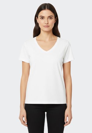 Femme aux cheveux noirs et raides, portant un T-shirt blanc uni à col en V et manches courtes, ainsi qu'un pantalon noir, debout devant un fond uni.